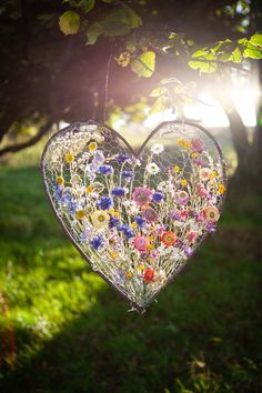 a heart shaped glass hanging from a tree in the sun with wildflowers on it