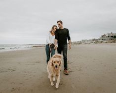 a man and woman walking their dog on the beach with houses in the back ground