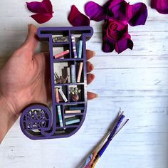 a hand holding a purple book shelf filled with books and pencils next to flowers