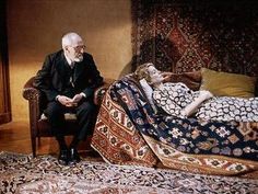 an old man sitting on a chair next to a woman laying on a bed in a room