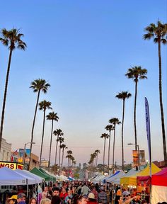 a crowd of people walking down a street next to tall palm trees and tents with signs on them