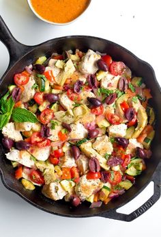 a skillet filled with vegetables and meat next to a bowl of tomato sauce on a white surface