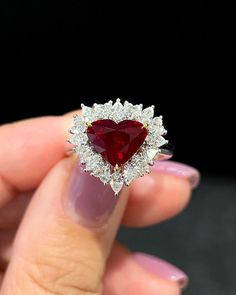 a woman's hand holding a heart shaped ring with diamonds around it and a red stone in the middle