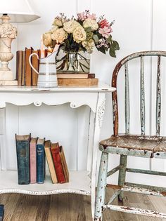 a white table with books and flowers on it next to a wooden chair in front of a lamp