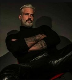 a man with grey hair and tattoos sitting on a red chair wearing black leather pants