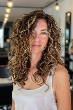 Woman with long, curly hair styled in beach waves, wearing a white tank top, smiling in a modern hair salon. Brown Honey Highlights, Honey Highlights