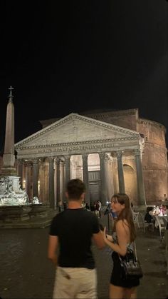 two people standing in front of an old building at night with the lights on and columns lit up