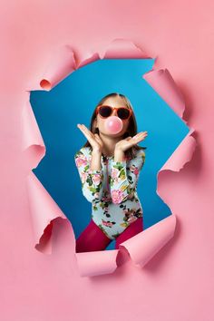 a woman wearing sunglasses and blowing bubble from her mouth through a hole in pink paper