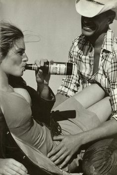 two women sitting on the ground drinking from bottles and talking to each other, with one woman wearing a cowboy hat