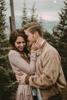 a man and woman embracing each other in front of pine trees at sunset with the sky behind them