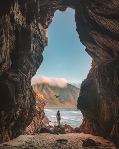 a man standing in the middle of a cave looking out at the ocean and mountains