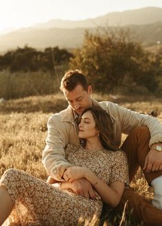 a man and woman cuddle together in the middle of a field with mountains in the background