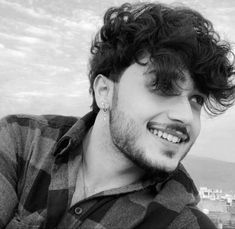 a man with curly hair smiling at the camera while wearing a checkered shirt and earrings