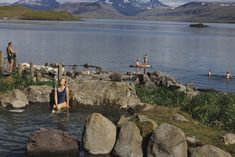 people are swimming in the water near some rocks and grass with mountains in the background