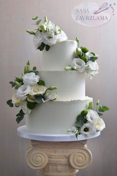 a three tiered cake with white flowers and greenery on the top is sitting on a pedestal