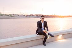 a man in a suit and tie sitting on a wall next to water holding a trumpet