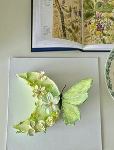 an open book on a table next to a plate with flowers and a butterfly sitting on it