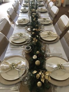 the table is set for christmas dinner with white and gold plates, silverware, and greenery