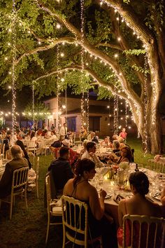 people are sitting at tables under trees with lights strung from the ceiling and on the ground