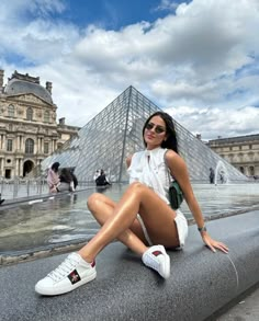 a woman sitting on the edge of a fountain in front of a large glass pyramid