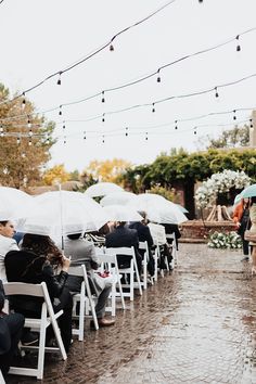 people sitting in chairs under umbrellas on a rainy day at an outdoor venue with string lights strung above them