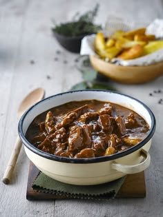 a bowl filled with beef and potatoes on top of a wooden table next to a spoon