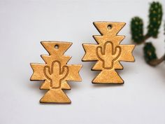 two brass colored metal charms with cactus designs on them and a plant in the background