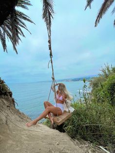 a woman sitting on a swing in the sand near the ocean with palm trees around her