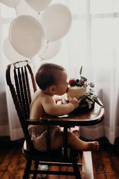 a baby sitting in a high chair with a cake on it's lap and balloons behind him