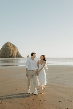 a man and woman standing on top of a sandy beach next to the ocean holding hands