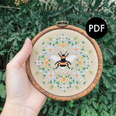 a hand holding up a small cross stitch bee in front of some green leaves and bushes