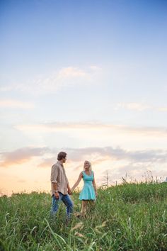 a man and woman holding hands while standing in tall grass at sunset with the sun setting behind them