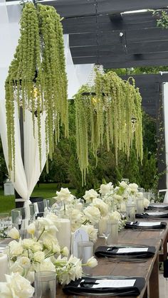 a long table with white flowers and greenery hanging from it's ceiling is set for an outdoor wedding reception