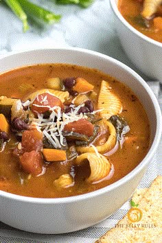 two white bowls filled with pasta and vegetable soup next to crackers on the side