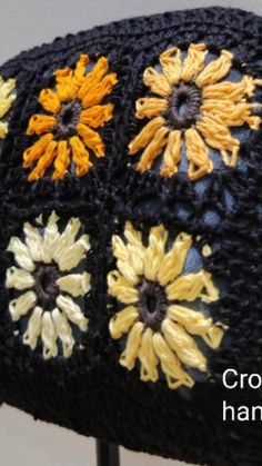 a black crocheted hat with yellow and orange flowers on the front, sitting on a metal stand