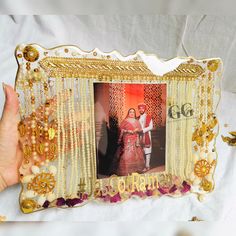 a person holding up a photo frame decorated with gold beads and flowers, in front of a white background