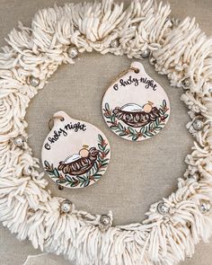 two ceramic ornaments with words on them sitting in front of a white wreath filled with tassels