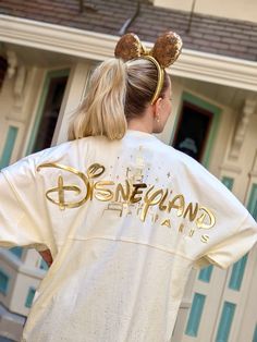 a woman wearing a white shirt with gold lettering on the front and back, standing in front of a house