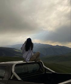 a woman sitting on the hood of a pickup truck looking out over rolling hills and mountains