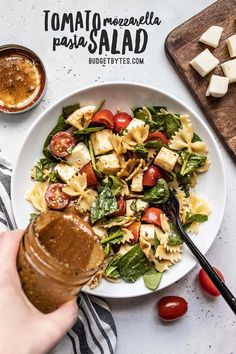 a person pouring dressing onto a plate of pasta salad with tomatoes, mozzarella and spinach