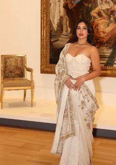 a woman standing in front of a painting wearing a white sari and gold jewelry