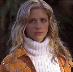 a woman with blonde hair wearing a yellow jacket and white turtle neck sweater looking at the camera