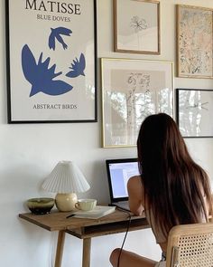 a woman sitting at a desk with a laptop computer in front of her and several framed pictures on the wall behind her