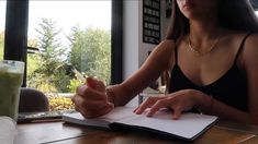 a woman sitting at a table writing on a book with a drink in the background