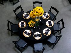 a round table with black chairs and yellow flowers in vases on the centerpiece