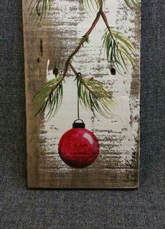 a red ornament hanging from a pine tree branch on a wooden plaque with white paint