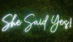 a neon sign that says she said yes on the side of a wall with green plants