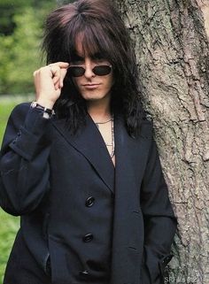 a man with long hair and sunglasses standing next to a tree wearing a suit jacket