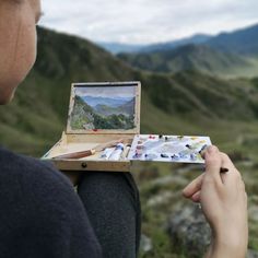 a woman is painting on an easel with mountains in the background and hills to the side