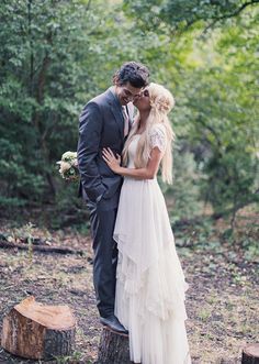 a man and woman in wedding clothes kissing on a tree stump with trees behind them